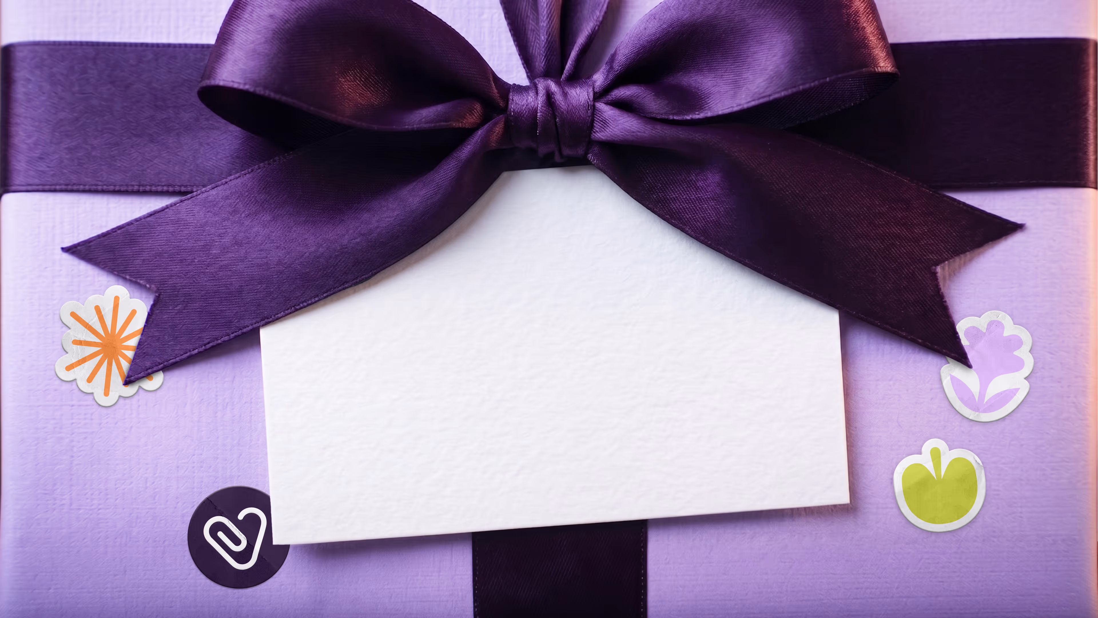 Caja de regalo color lavanda con lazo morado satinado y tarjeta blanca en el centro, decorada con pegatinas coloridas.