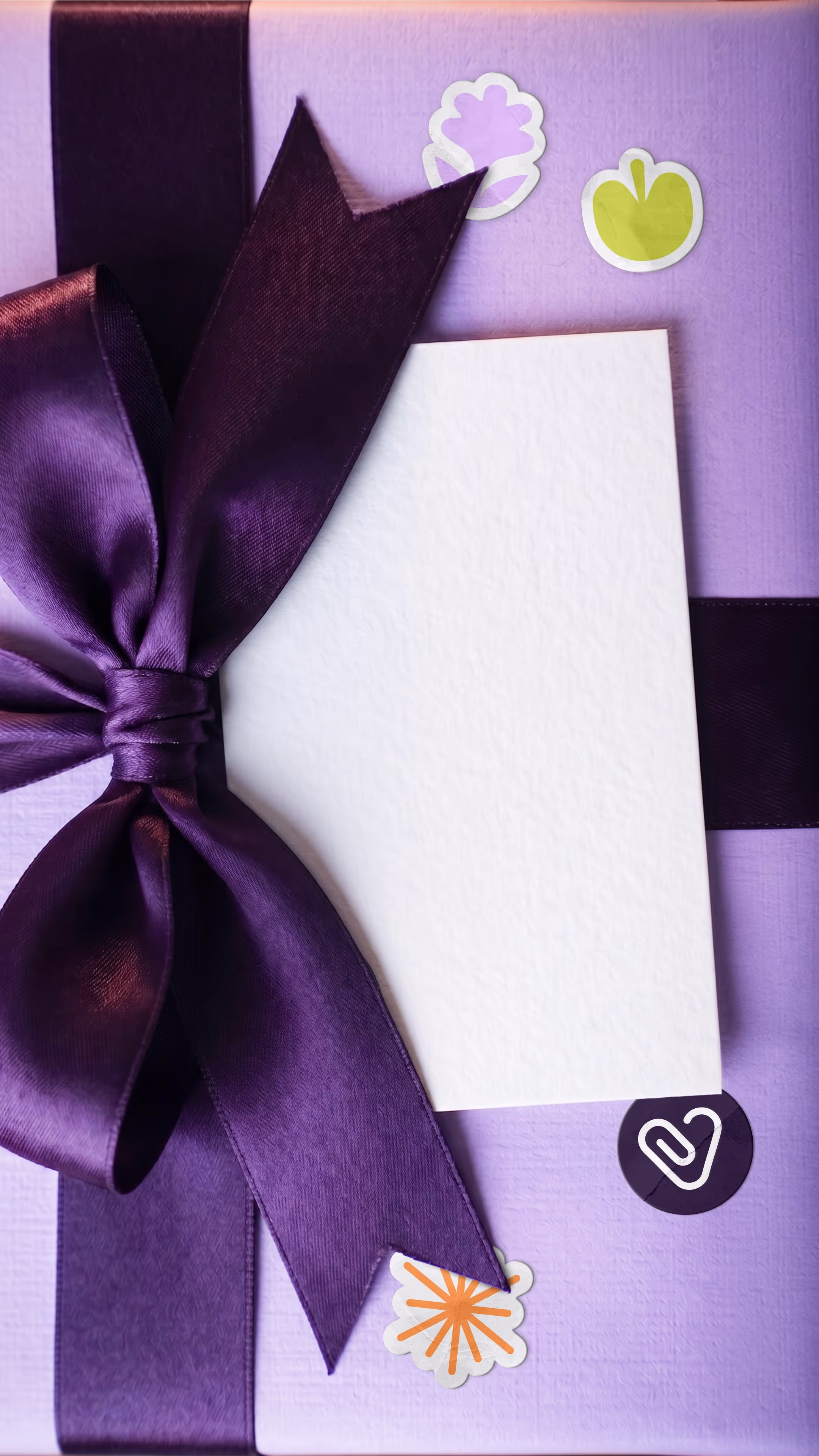 Caja de regalo color lavanda con lazo morado satinado y tarjeta blanca en el centro, decorada con pegatinas coloridas.